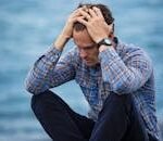 A man in a plaid shirt sits by the water looking distressed, symbolizing stress.